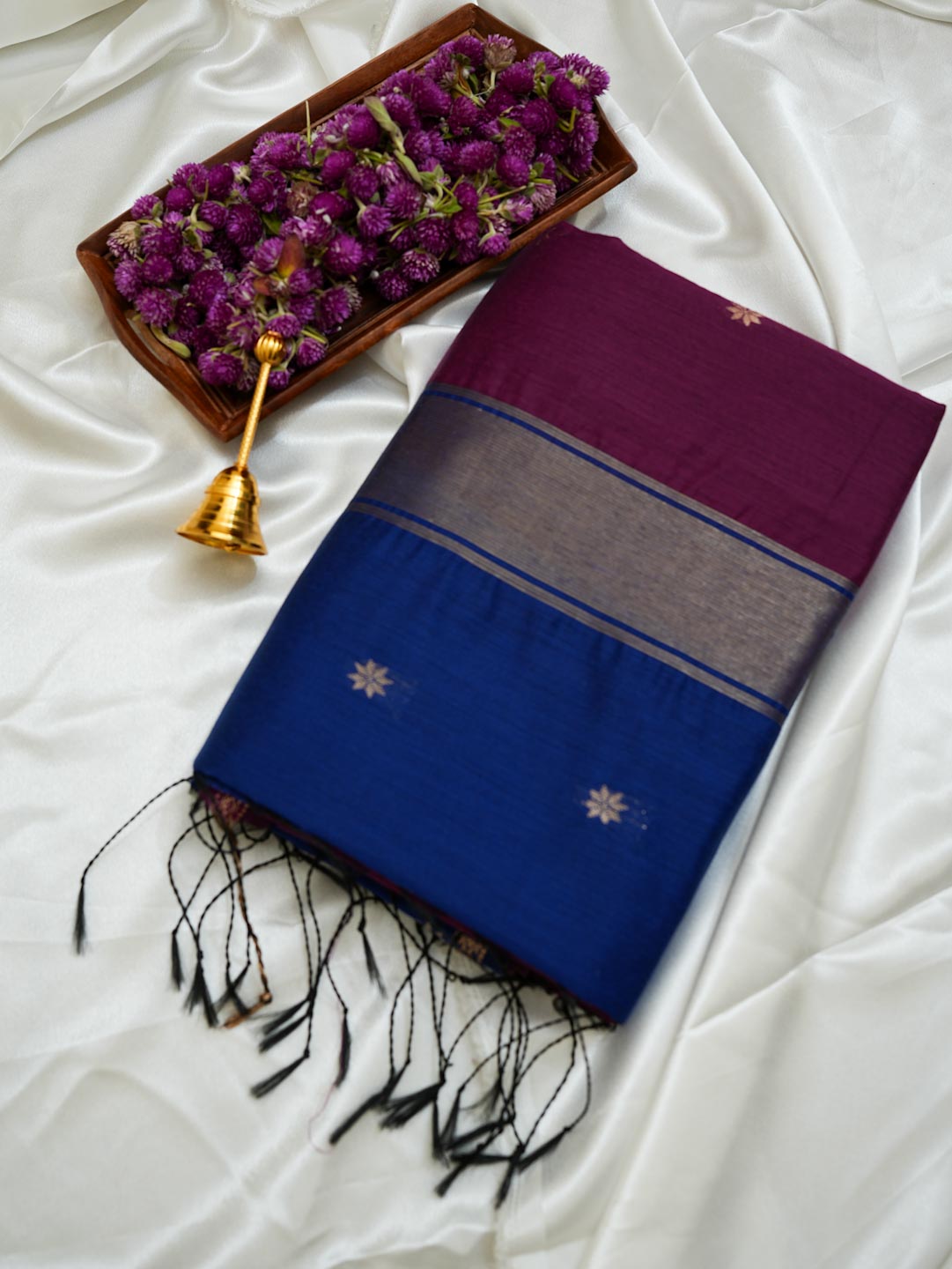 A folded purple and blue saree with black fringe, displayed on a white satin fabric alongside a tray of purple flowers and a small gold bell.