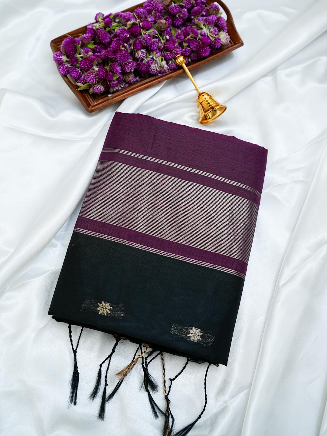 A folded dark purple and black saree with metallic silver stripes and gold zari embroidery, displayed on a white satin fabric alongside a small tray of purple flowers and a gold bell.